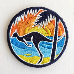Può includere: Una toppa rotonda ricamata con una silhouette di canguro nera che salta davanti a un tramonto sul mare blu. La toppa ha un contorno bianco ed è su uno sfondo blu scuro.