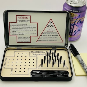 May include: A black metal box with a wooden game board inside. The game board has two sections, one for a solitaire game and one for a pyramid game. The box also includes a set of black pegs and instructions for both games.