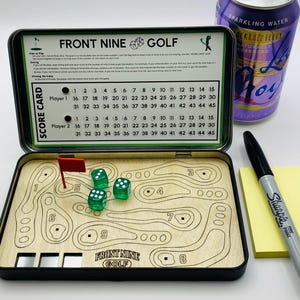 May include: A Front Nine Golf game set in a black tin. The game board is made of wood and features a golf course design. Includes green dice, a red flag, and a score card. A can of sparkling water and a black marker are also present.
