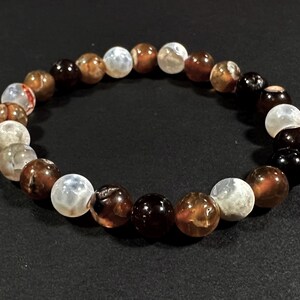 May include: A beaded bracelet featuring a variety of earth-toned beads. The beads are primarily brown, white, and black, with variations in color and pattern. The bracelet is a circular shape, suitable for wrist wear.