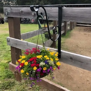 May include: A black metal hanging planter bracket attached to a wooden fence with a colorful flower basket hanging from it.
