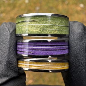 May include: Three stacked rings with a silver band. The top ring is green, the middle ring is purple, and the bottom ring is yellow. Each ring has a textured, layered design with white lines.
