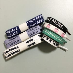 Puede incluir: Una colección de cordones de zapatos coloridos con varios diseños impresos, que incluyen "RESALE", "LUCKY 13", "SAY MORE", "Anniversary" y "The TSA's Finest".