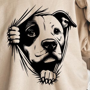 Puede incluir: Una sudadera beige con una ilustración en blanco y negro de la cabeza de un perro asomándose por un agujero rasgado en la tela.