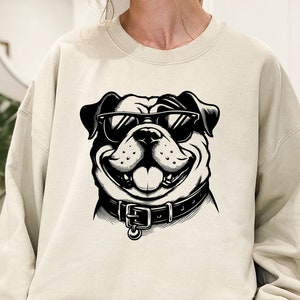May include: A cream-colored sweatshirt with a black and white illustration of a bulldog wearing sunglasses. The bulldog is smiling and has a collar.