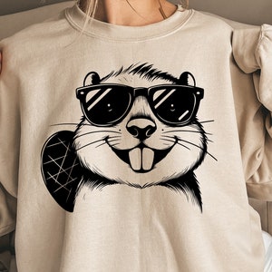 May include: Beige sweatshirt featuring a black and white graphic of a beaver wearing sunglasses. The beaver is smiling, and the design includes a beaver tail. The sweatshirt is a neutral color and has a crew neck.