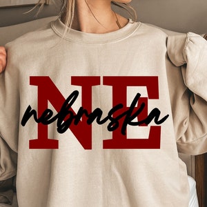 May include: A beige sweatshirt with the letters "NE" in a bold, red font, with "nebraska" written in a cursive black font across the letters. The sweatshirt is a crew neck style.