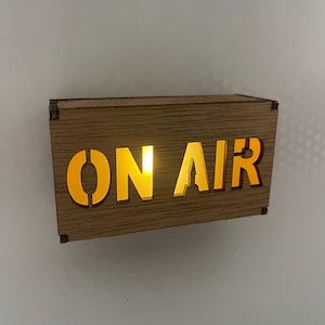 May include: A wooden "ON AIR" sign with illuminated yellow lettering. The sign is rectangular and has a natural wood grain finish. The letters are cut out, allowing the light to shine through, creating a warm glow.