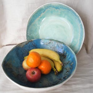 May include: Two ceramic bowls, one containing fruit. The larger bowl is a deep blue with a white rim, holding bananas, oranges, and an apple. A second, similar bowl is in the background, with a light blue and white colour scheme. The bowls are on a white surface.