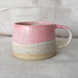 Könnte beinhalten: Eine handgefertigte Keramik-Tasse mit rosa Oberteil, weißem Mittelteil und gesprenkelter beiger Basis. Die Tasse hat einen gebogenen Henkel und ist für heiße oder kalte Getränke geeignet. Die Tasse ist etwa 9 cm hoch und 10 cm breit.