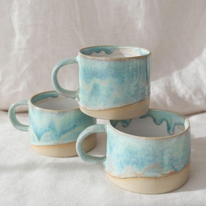 May include: Three ceramic mugs with a light blue glaze and a tan base are stacked. The mugs have a simple handle and a smooth, glossy finish. The mugs are handmade and have a unique, organic shape. The mugs are suitable for tea or coffee.