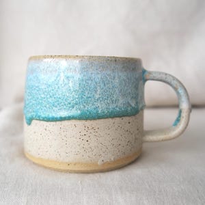 May include: A ceramic mug with a speckled cream base and a turquoise blue top. The mug features a rounded handle and a textured, glazed finish. The mug is suitable for hot drinks.