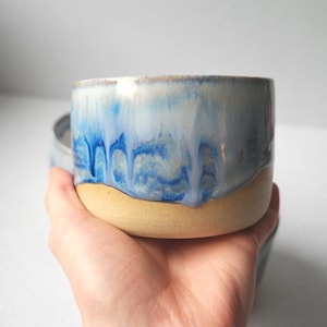 May include: A ceramic mug with a cylindrical shape, featuring a smooth, light beige unglazed base and a glossy, drippy blue and white glaze on the upper portion. The glaze creates a layered, flowing effect that transitions from white to deep blue.