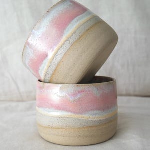 May include: Two handmade ceramic cups stacked together. The cups have a speckled, beige base with bands of light pink, white, and yellow glaze. The top cup is tilted, revealing the interior. The cups are likely used for drinking beverages.