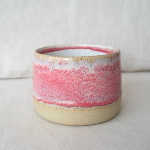 Puede incluir: Una taza de cerámica hecha a mano con un vibrante esmalte rojo y blanco. La taza tiene una base beige y un interior blanco. El esmalte rojo tiene un aspecto texturizado y moteado. La taza es cilíndrica y parece de tamaño pequeño.