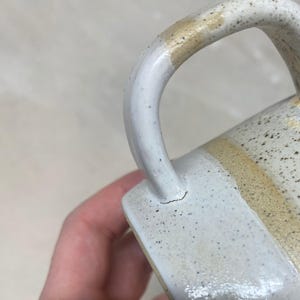 May include: Close-up of a handmade ceramic mug with a speckled white glaze. The handle is white with a tan accent. The mug has a crack near the base of the handle. The mug is a neutral color and has a rustic aesthetic.