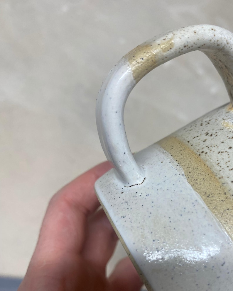 May include: Close-up of a handmade ceramic mug with a speckled white glaze. The handle is white with a tan accent. The mug has a crack near the base of the handle. The mug is a neutral color and has a rustic aesthetic.
