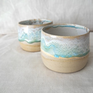 May include: Two handmade ceramic cups with a coastal design. The cups have a beige base, with a white, turquoise, and green glaze that resembles ocean waves. The rims are slightly irregular.