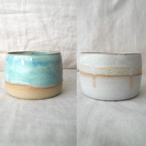 Op de afbeelding: Twee handgemaakte keramische bekers. Eén is lichtblauw met een beige basis, de andere is wit met een beige streep en druppels. Beide hebben een cilindrische vorm en worden waarschijnlijk gebruikt om uit te drinken.