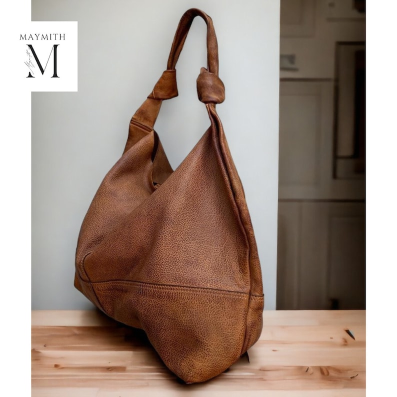 May include: A brown leather shoulder bag with a knotted handle. The bag has a textured surface and a large capacity. The bag is sitting on a wooden surface. The logo "MAYMITH" is in the upper left corner.