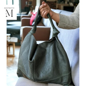 May include: A dark green leather shoulder bag with a knotted handle. The bag has a slouchy design and visible stitching details. The bag is held by a person.