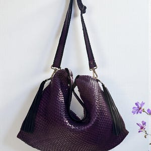 K&ouml;nnte beinhalten: Eine tiefviolette, gewebte Handtasche mit Schulterriemen und zwei schwarzen Quasten. Die Tasche h&auml;ngt vor einer wei&szlig;en Wand. Der Riemen ist dunkelviolett mit schwarzem Besatz und silberfarbener Hardware.