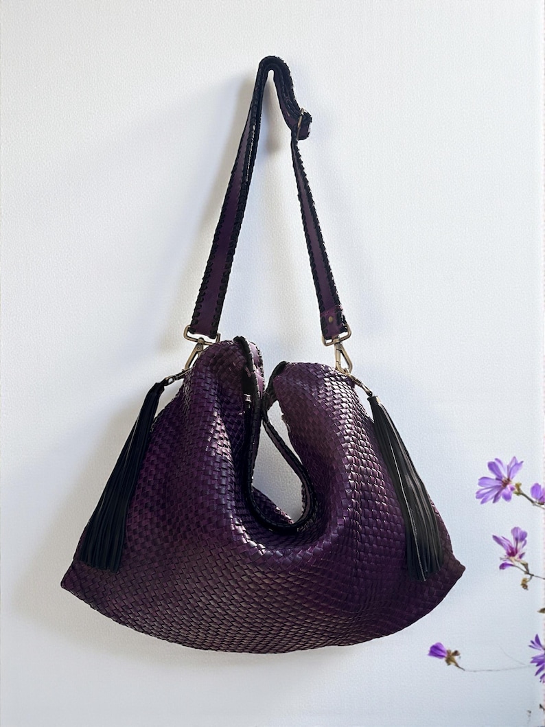 K&ouml;nnte beinhalten: Eine tiefviolette, gewebte Handtasche mit Schulterriemen und zwei schwarzen Quasten. Die Tasche h&auml;ngt vor einer wei&szlig;en Wand. Der Riemen ist dunkelviolett mit schwarzem Besatz und silberfarbener Hardware.