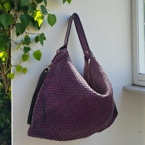 K&ouml;nnte beinhalten: Eine gewebte lila Umh&auml;ngetasche mit langem Riemen und schwarzer Quaste. Die Tasche h&auml;ngt an einer wei&szlig;en Wand, mit gr&uuml;nen Bl&auml;ttern im Hintergrund. Die Tasche hat einen Rei&szlig;verschluss.