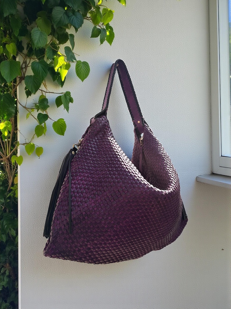 K&ouml;nnte beinhalten: Eine gewebte lila Umh&auml;ngetasche mit langem Riemen und schwarzer Quaste. Die Tasche h&auml;ngt an einer wei&szlig;en Wand, mit gr&uuml;nen Bl&auml;ttern im Hintergrund. Die Tasche hat einen Rei&szlig;verschluss.