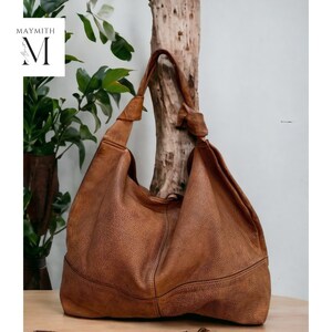 May include: A large, brown leather hobo bag with a textured surface. The bag has a single shoulder strap and a slouchy design. The bag is displayed on a wooden surface with greenery in the background. The brand name "MAYMITH" is in the upper left corner.
