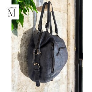 K&ouml;nnte beinhalten: Eine dunkelblaue Lederhandtasche mit zwei Henkeln und einem Rei&szlig;verschluss. Die Tasche hat eine strukturierte Oberfl&auml;che und eine kleine Tasche an der Seite. Die Tasche h&auml;ngt an einer Holzstange vor einer Steinmauer. Der Markenname "MAYMITH" ist sichtbar.