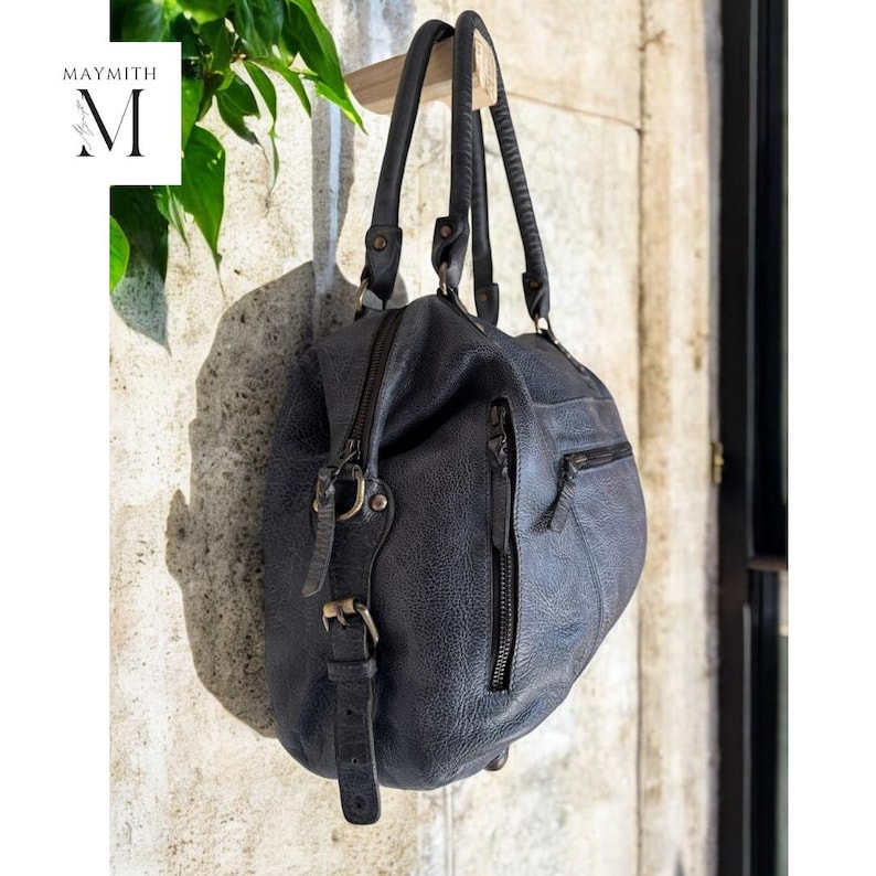 K&ouml;nnte beinhalten: Eine dunkelblaue Lederhandtasche mit zwei Henkeln und einem Rei&szlig;verschluss. Die Tasche hat eine strukturierte Oberfl&auml;che und eine kleine Tasche an der Seite. Die Tasche h&auml;ngt an einer Holzstange vor einer Steinmauer. Der Markenname "MAYMITH" ist sichtbar.