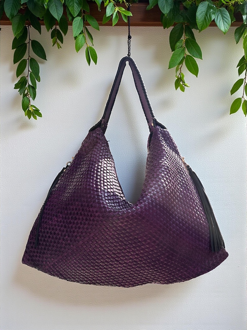 K&ouml;nnte beinhalten: Eine gewebte, lila Umh&auml;ngetasche mit einem gro&szlig;en, l&auml;ssigen Design. Die Tasche hat ein strukturiertes Muster und lange, dunkle Griffe. Schwarze Quasten h&auml;ngen an den Seiten. Die Tasche h&auml;ngt vor einem wei&szlig;en Hintergrund, mit gr&uuml;nen Bl&auml;ttern im Hintergrund.