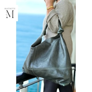 May include: A dark green leather shoulder bag with a knotted strap. The bag has a slouchy design and visible stitching. The logo "MAYMITH" is in the upper left corner. The background is a blurred view of the ocean.
