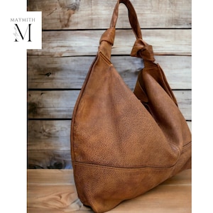 May include: A brown leather shoulder bag with a knotted strap. The bag has a textured surface and a large, slouchy design. The bag is set against a wooden background, with the brand name "MAYMITH" in the upper left corner.