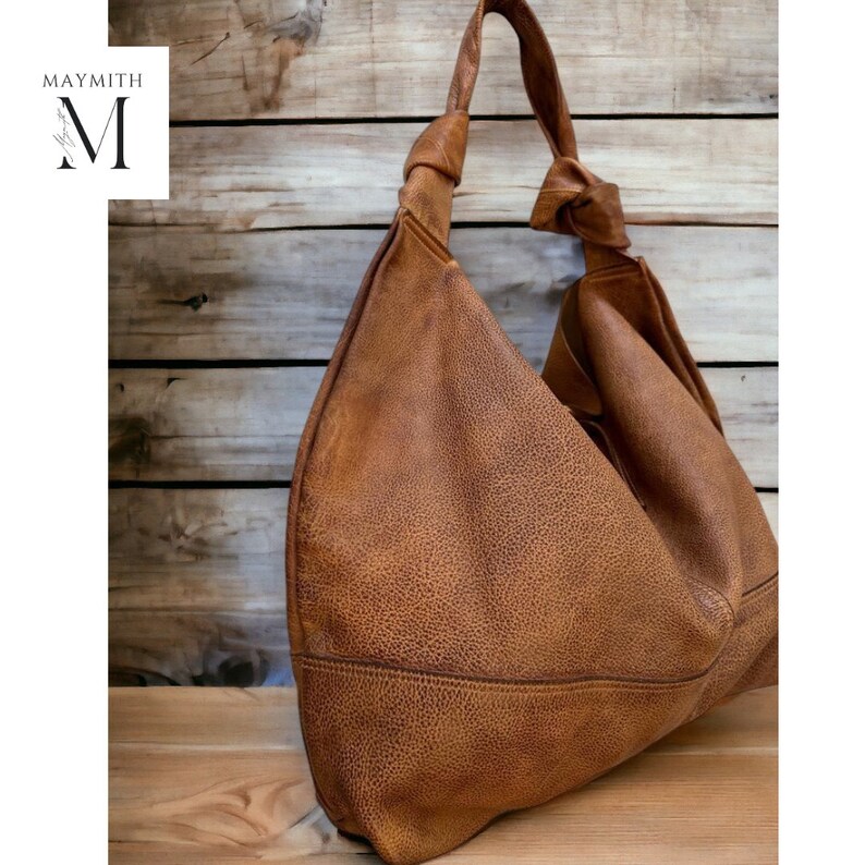 May include: A brown leather shoulder bag with a knotted strap. The bag has a textured surface and a large, slouchy design. The bag is set against a wooden background, with the brand name "MAYMITH" in the upper left corner.