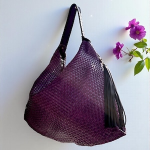 K&ouml;nnte beinhalten: Eine gewebte, auberginefarbene Umh&auml;ngetasche mit schwarzem Riemen und Quaste. Die Tasche hat ein gro&szlig;es, l&auml;ssiges Design und ein strukturiertes Aussehen. Der Riemen ist mit einer Metallniete an der Tasche befestigt. Die Tasche h&auml;ngt vor einem wei&szlig;en Hintergrund.