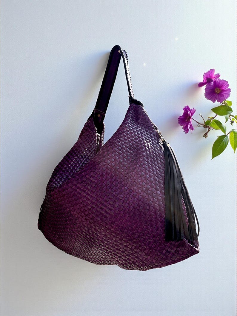 K&ouml;nnte beinhalten: Eine gewebte, auberginefarbene Umh&auml;ngetasche mit schwarzem Riemen und Quaste. Die Tasche hat ein gro&szlig;es, l&auml;ssiges Design und ein strukturiertes Aussehen. Der Riemen ist mit einer Metallniete an der Tasche befestigt. Die Tasche h&auml;ngt vor einem wei&szlig;en Hintergrund.