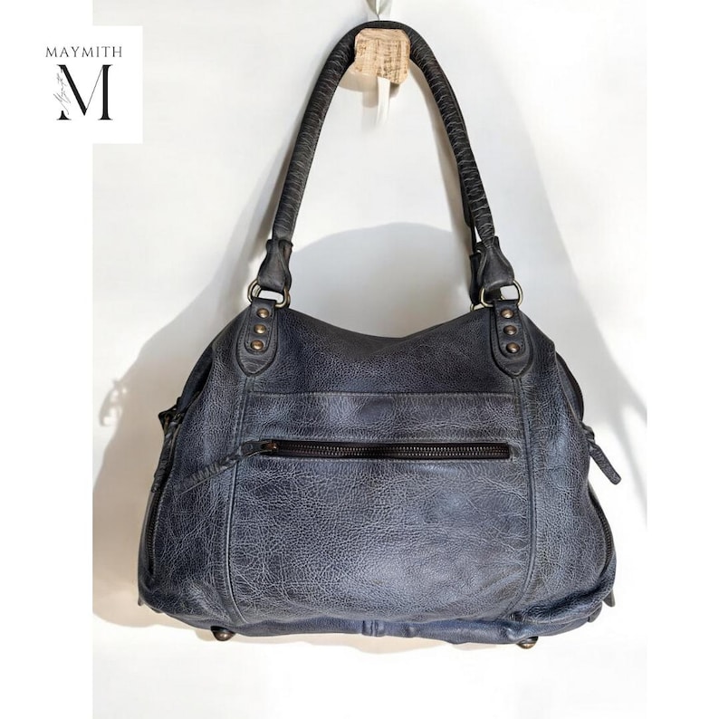 K&ouml;nnte beinhalten: Eine blaue Handtasche aus Leder mit Used-Look, mit einem oberen Griff und einer Rei&szlig;verschlusstasche. Die Tasche hat eine strukturierte Optik und Metallbeschl&auml;ge. Der Markenname "MAYMITH" und der Buchstabe "M" sind auf wei&szlig;em Hintergrund zu sehen.