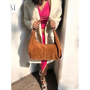 Puede incluir: Un bolso de mano de ante marrón con una correa larga. El bolso lo lleva una persona que lleva un abrigo blanco y una blusa rosa.