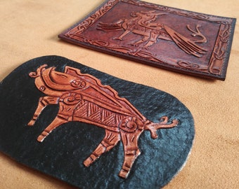 Op de afbeelding: Twee leren patches met gegraveerde dierenontwerpen. De eerste patch is bruin en heeft een gevleugeld wezen met een lange staart. De tweede patch is zwart en heeft een stier met ingewikkelde details.