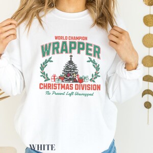 May include: White sweatshirt with a Christmas-themed graphic that says "World Champion Wrapper Christmas Division No Present Left Unwrapped". The graphic features a green Christmas tree with red and green presents, red berries, and a gold star on top.