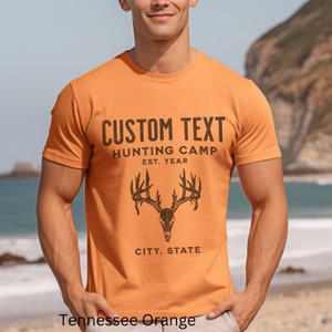 May include: Orange t-shirt with the text "CUSTOM TEXT HUNTING CAMP EST. YEAR CITY, STATE" and a graphic of a deer skull with antlers.