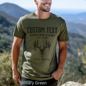 May include: A military green t-shirt with a black deer skull graphic and the text "CUSTOM TEXT HUNTING CAMP EST. YEAR CITY, STATE".