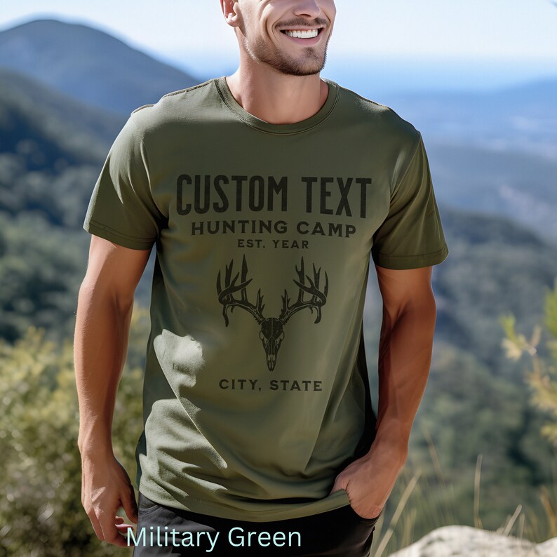 May include: A military green t-shirt with a black deer skull graphic and the text "CUSTOM TEXT HUNTING CAMP EST. YEAR CITY, STATE".