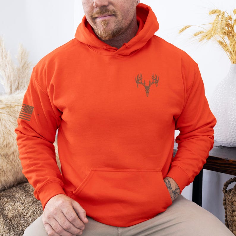 May include: Bright orange hoodie with a front pocket and a drawstring hood. The hoodie features a deer antler graphic on the chest and a small American flag on the left sleeve. The hoodie is made of a soft material.