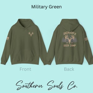 May include: Military green hoodie with a front pocket and a hood. The front features a deer antler graphic, and the back displays "Jackson's Deer Camp" with a deer antler graphic and the text "ESTD 1984". The sleeve has a small American flag.