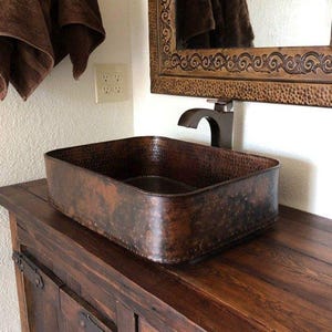 May include: A rustic wooden vanity with a hammered copper sink and a matching ornate mirror. The vanity has a distressed wood finish and features iron hardware.