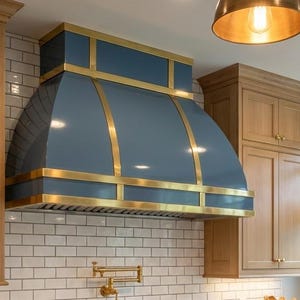May include: A custom kitchen range hood in a glossy blue finish with gold-colored trim. The hood is mounted above a white tile backsplash and is flanked by light wood cabinets. A brass faucet is visible.
