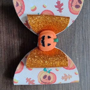 May include: A white and orange bow with a pumpkin design. The bow is made of fabric and has a glitter finish. The pumpkin is a 3D embellishment.